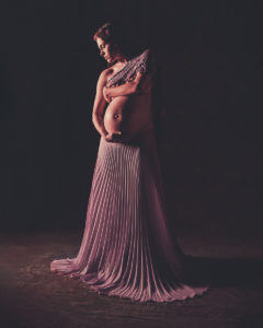 Artistic nude of pregnant woman in long rose colored pleated skirt covering her chest with her arm and rose color shawl