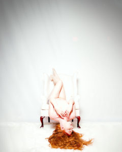 Artistic nude of redhead upside down on white wingback chair in white room with hair fanned out white fur