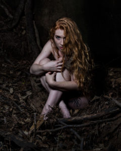 Artistic nude of nude redheaded woman huddled with arms and legs crossed on the forest floor