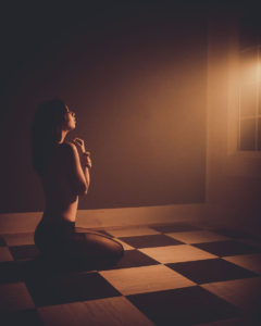 Artistic nude of woman kneeling in front of window with arms crossed in front of her chest bathed in yellow tinted light