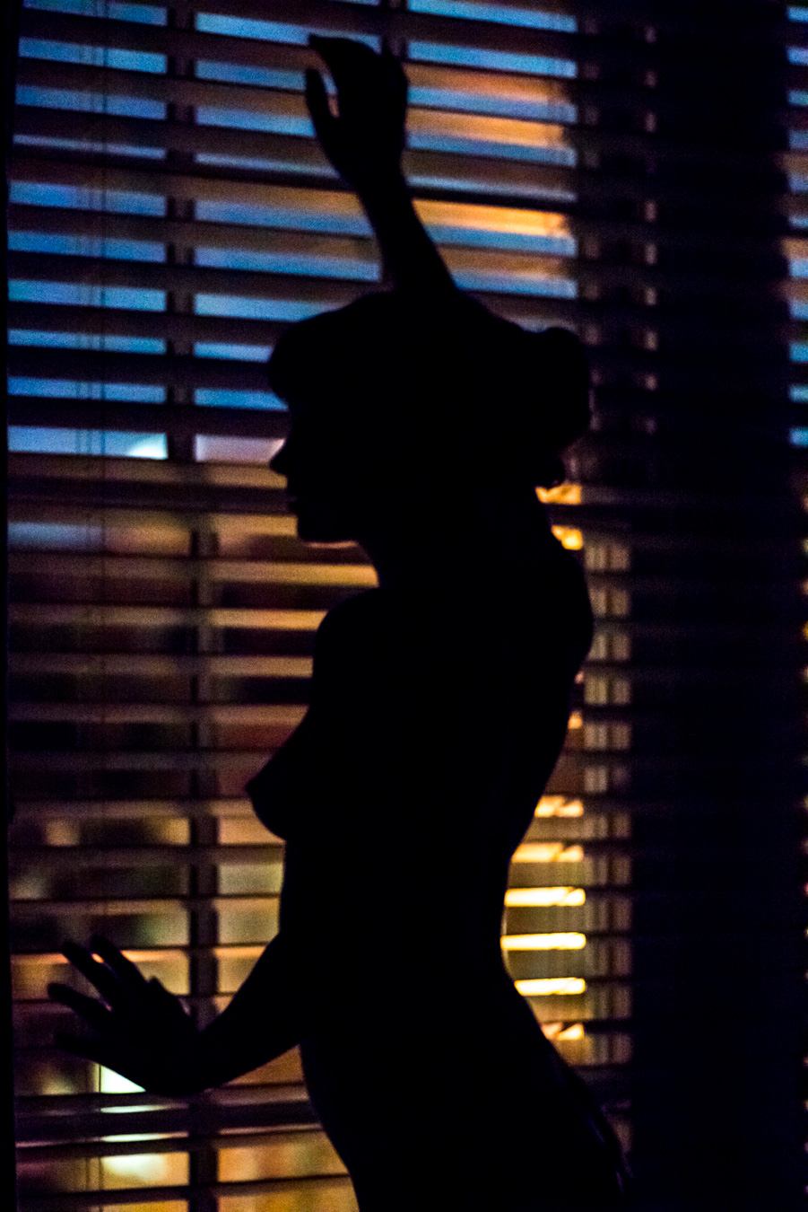 Silhouette of a person standing near window blinds at night, with colorful city lights outside creating a striking image.