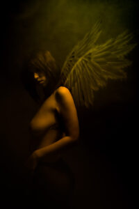 A nude woman with wings posing in a dark room.