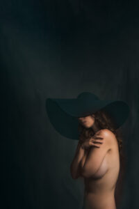 A nude woman in a black hat posing in front of a dark background.