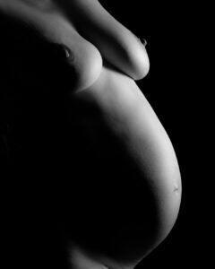 A black and white side profile image of a pregnant person's bare torso and chest against a dark background.