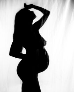 Silhouette of a pregnant woman posing with one hand on her hip and the other hand raised above her head against a bright background.