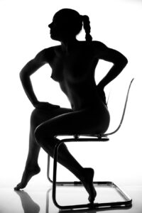 Silhouette of a nude person with braided hair sitting sideways on a transparent chair against a white background.