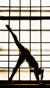 A person performs a yoga pose, balancing on hands and one foot with the other leg extended upward, silhouetted against a large grid window.