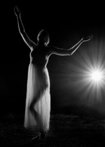 A person in a sheer dress poses gracefully with arms raised, backlit by a bright light source in a dark setting.