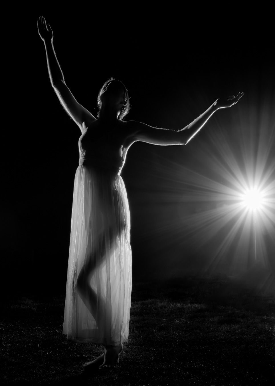 A person in a sheer dress poses gracefully with arms raised, backlit by a bright light source in a dark setting.