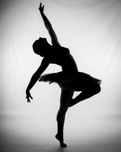 Silhouette of a ballet dancer in a tutu, posed on one leg with arms extended, against a white background.