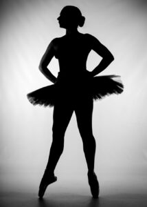 Silhouette of a ballet dancer standing en pointe with hands on hips, wearing a tutu against a plain, light background.