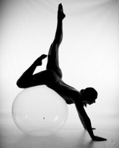 A person poses nude in an athletic position, balancing on a large exercise ball with one leg extended upward and both hands on the floor, set against a white background.