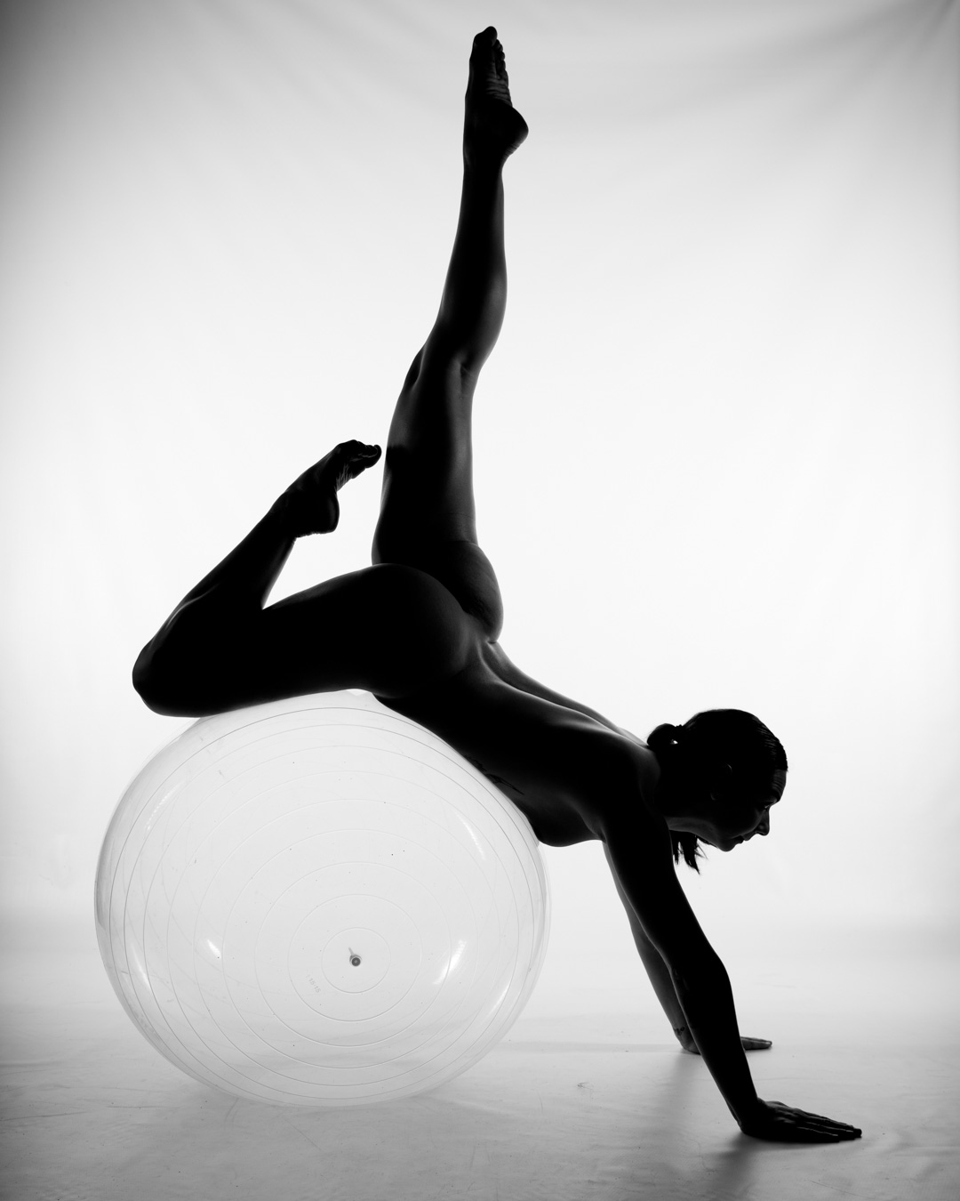 A person poses nude in an athletic position, balancing on a large exercise ball with one leg extended upward and both hands on the floor, set against a white background.