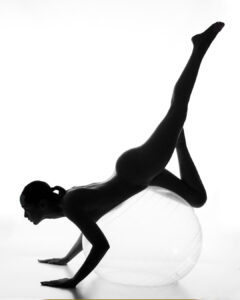 A nude person balances on a large exercise ball, supporting their body with their hands and one leg while the other leg is extended upward.