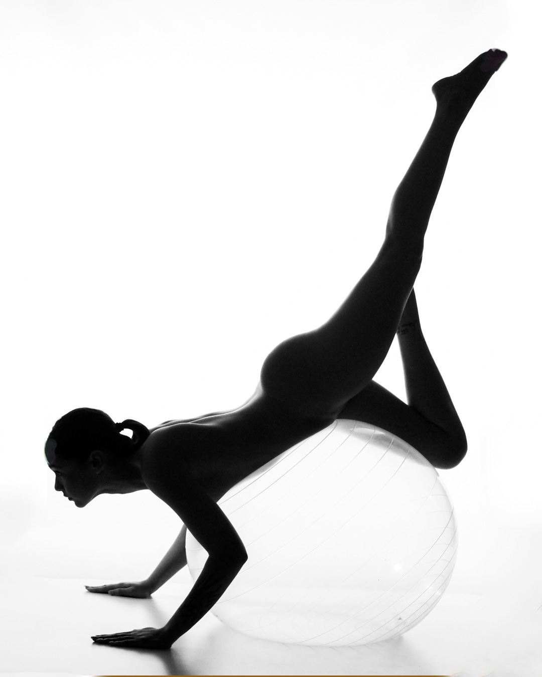 A nude person balances on a large exercise ball, supporting their body with their hands and one leg while the other leg is extended upward.