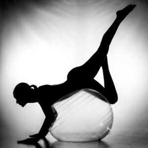 A person in silhouette balances on an exercise ball with one leg bent and the other extended upward against a plain background.