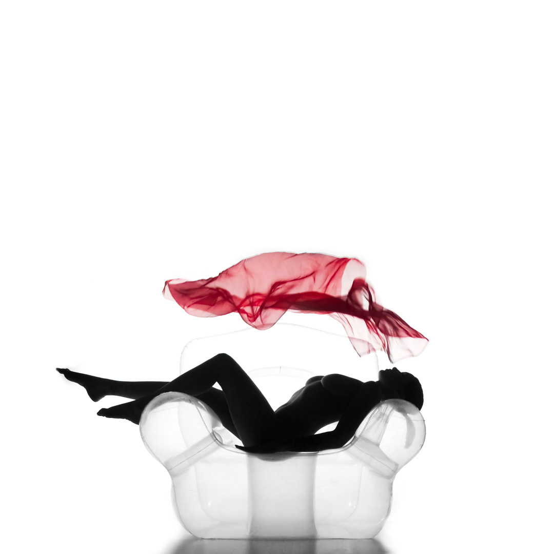 Silhouette of a person reclining on a transparent chair with a red sheer fabric floating above their head against a white background.