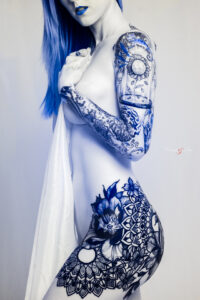 A woman with blue hair and lips displays intricate blue floral tattoos on her arm, shoulder, and hip, holding a white cloth in front of her body.